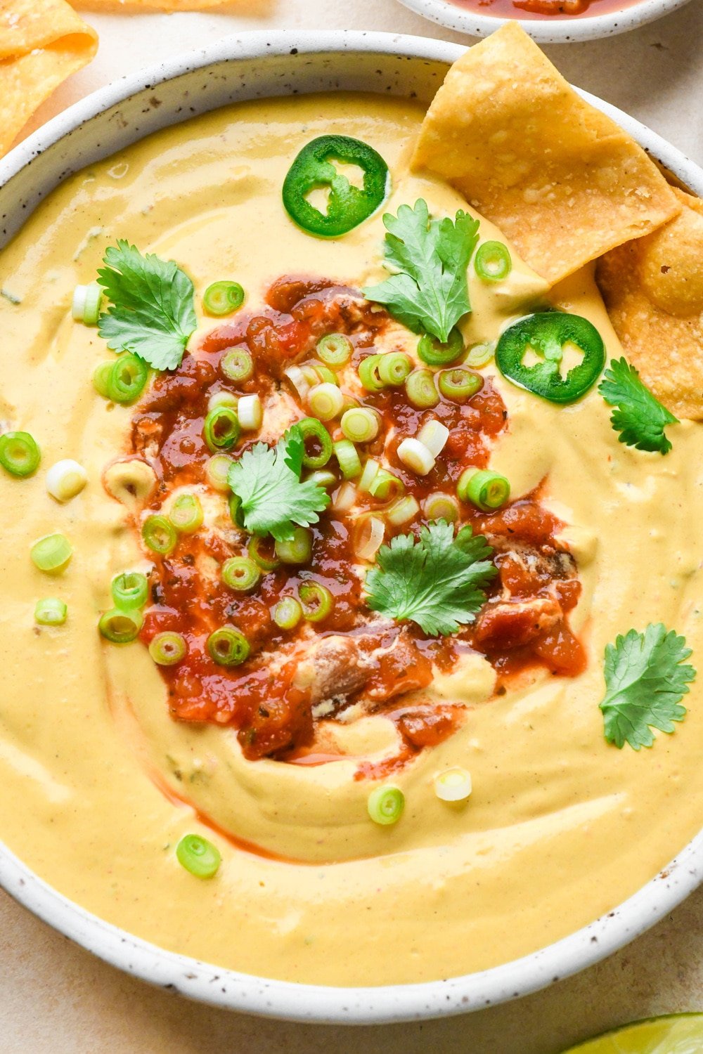 A medium sized shallow ceramic bowl filled with creamy cashew queso that is topped with a swirl of salsa, green onions, cilantro, and thinly sliced jalapeño. There are several chips dipped into the bowl to show the creamy texture.