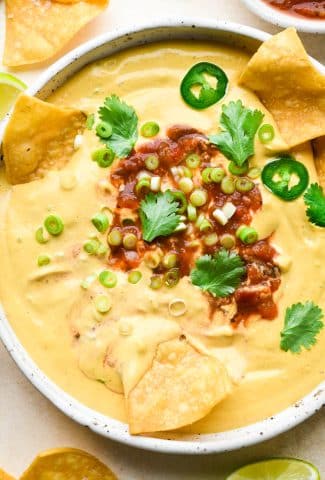 A medium sized shallow ceramic speckled bowl filled with creamy cashew queso that is topped with a swirl of salsa, green onions, cilantro, and thinly sliced jalapeño. There are several chips dipped into the bowl to show the creamy texture.