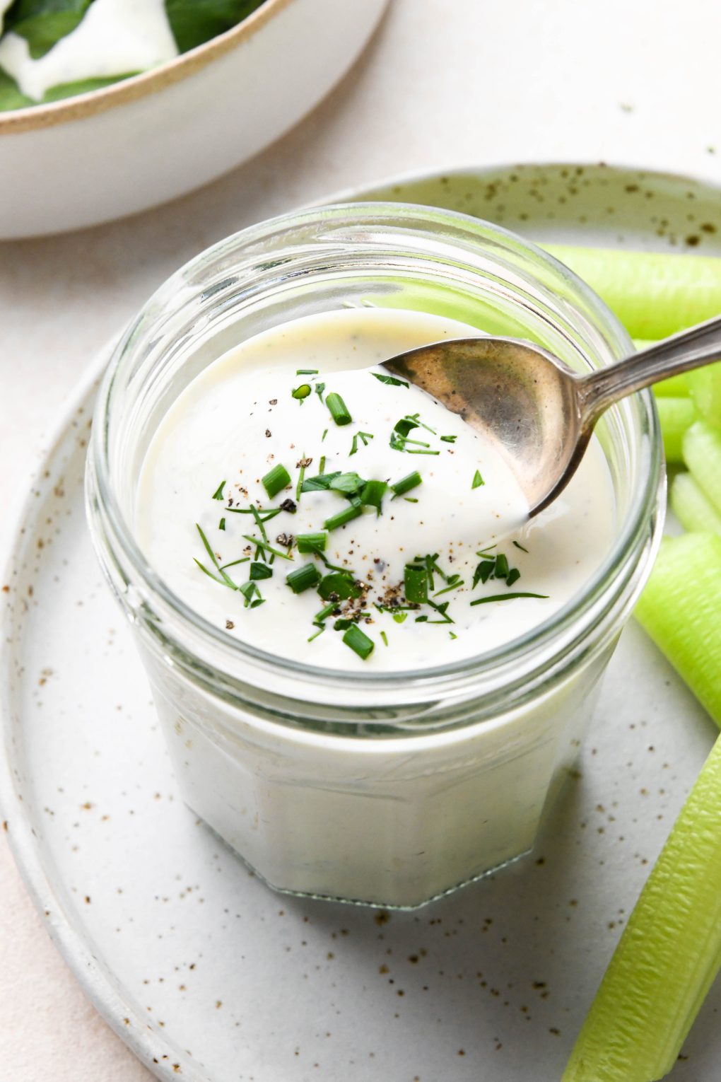 Dairy free ranch dressing in a small glass jar next to celery sticks with a spoon dipping into the dressing. On a cream colored background.