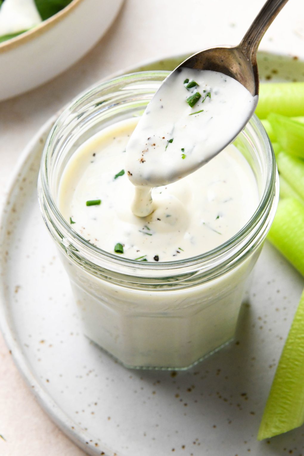 Dairy free ranch dressing in a small glass jar next to celery sticks with a spoon drizzling some of the dressing. On a cream colored background.