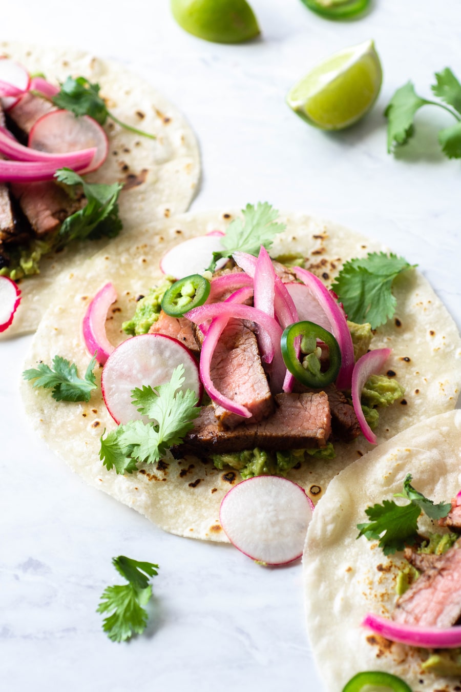 Side angle of multiple steak tacos on a white background with cilantro, thinly sliced jalapeno, pickled red onions