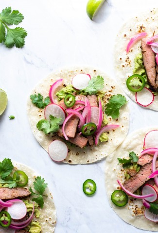 Multiple steak tacos on a white background with cilantro, thinly sliced jalapeno, pickled red onions
