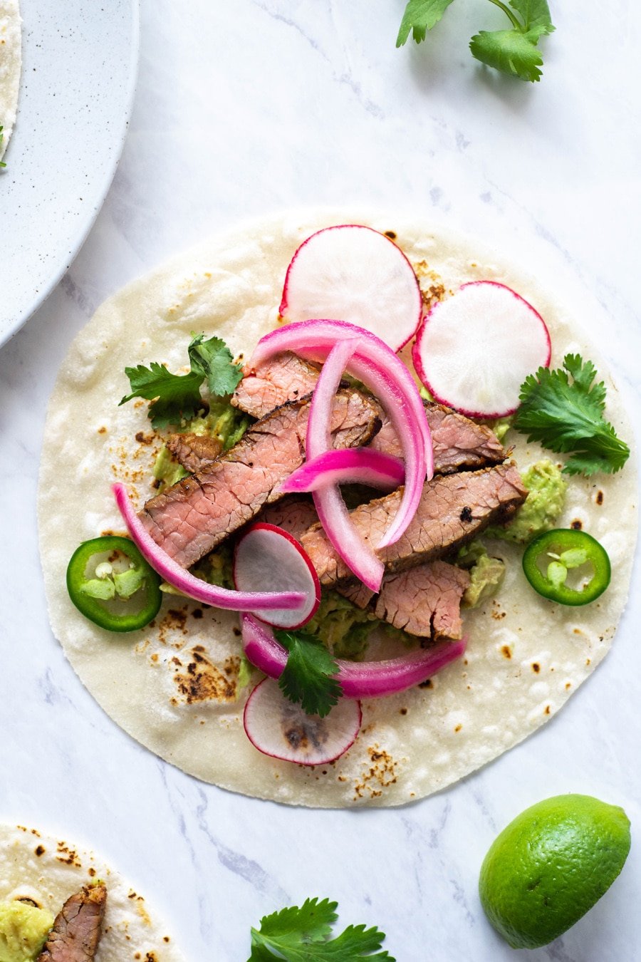 A close up shot of a steak taco on a white background with cilantro, thinly sliced jalapeno, pickled red onions