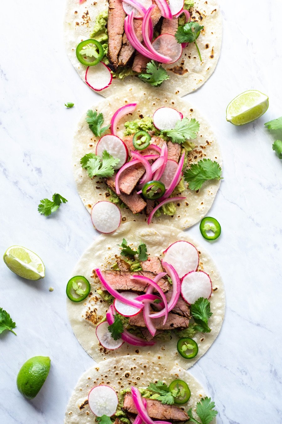 Multiple steak tacos lined up from the top to the bottom of the image on a white background with cilantro, thinly sliced jalapeno, pickled red onions