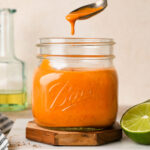 Creamy bright orange roasted red pepper dressing in a short, square, 16-ounce mason jar next to a few lime wedges and a skinny light green bottle of olive oil. A spoon has dipped into the dressing and is lifted out of the jar, a little dressing dripping back into the jar to show the velvety texture.