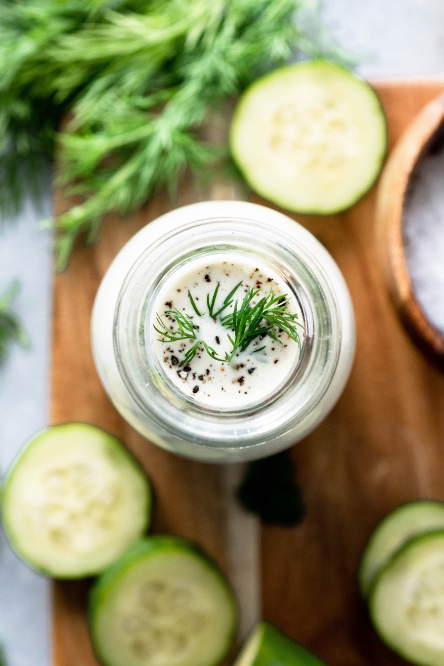 Overhead view of a jar of homemade dairy free ranch with herbs and cucumbers on a wooden board