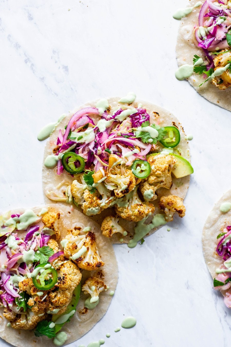 Overhead shot of layered mexican roasted cauliflower tacos with cabbage slat, sliced jalapenos, and a drizzle of cashew lime crema