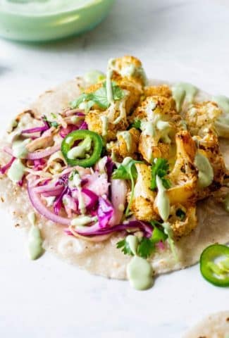 Side angle view of mexican roasted cauliflower tacos with cabbage slat, sliced jalapenos, and a drizzle of cashew lime crema on a white background