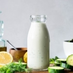 Straight on view of a jar of homemade dairy free ranch with lemon, herbs, and cucumbers on a wooden board
