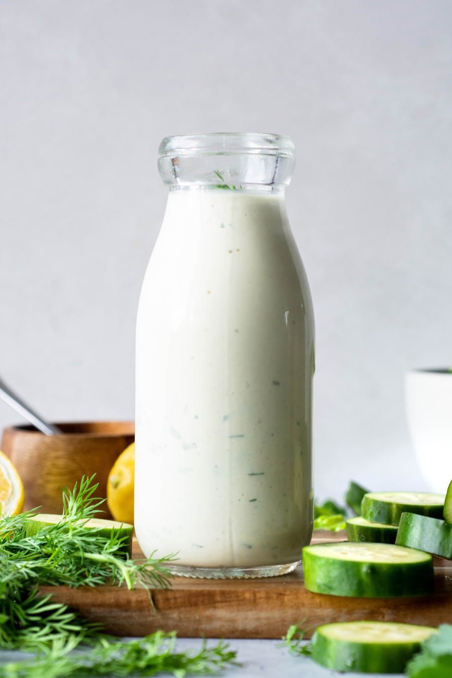 Straight on view of a jar of homemade dairy free ranch with lemon, herbs, and cucumbers on a wooden board
