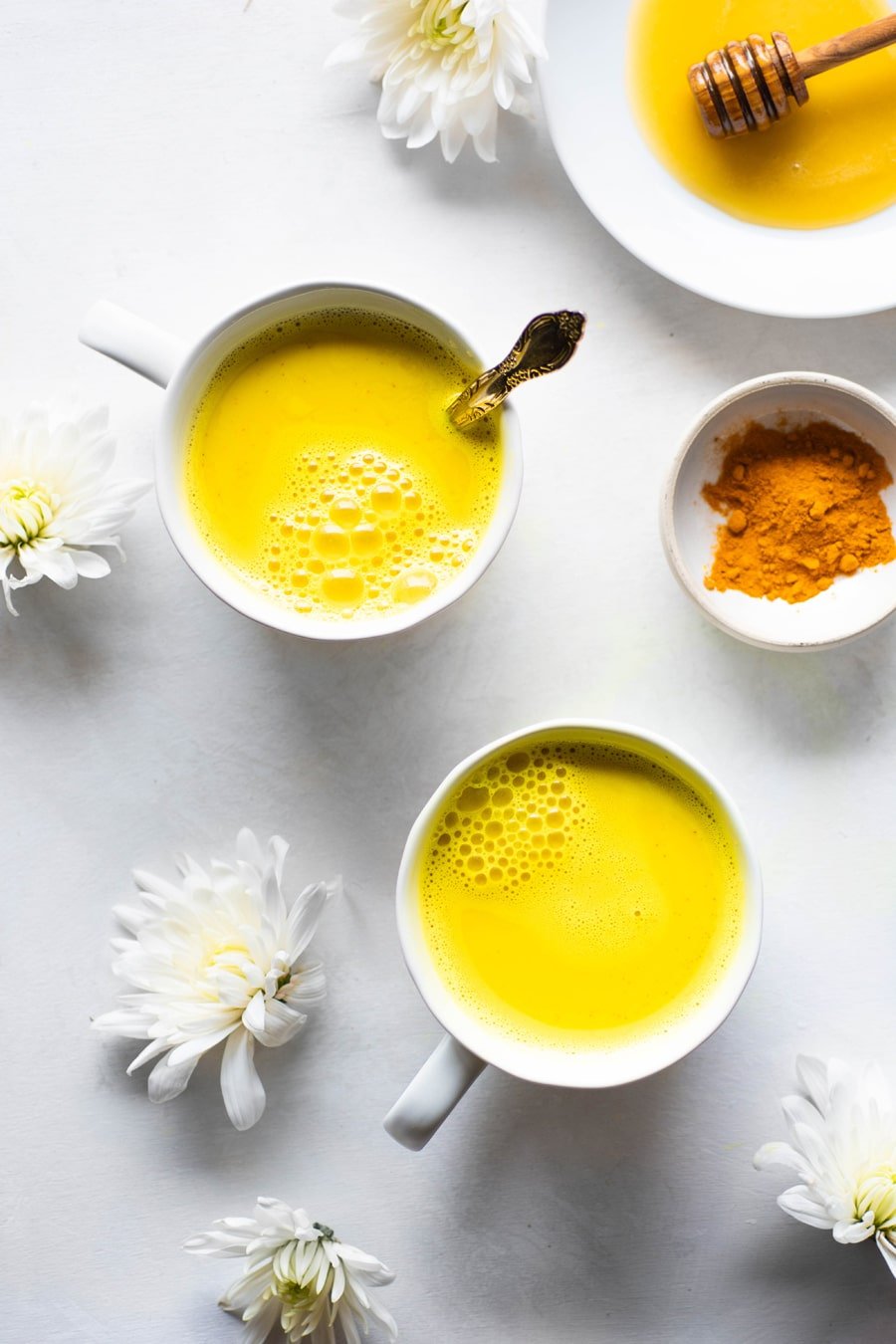 Two bright yellow golden milk latte in a white mug with white flowers and a small bowl of turmeric