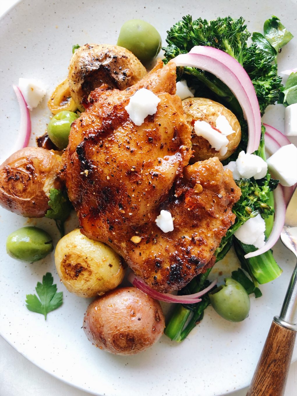 A close up view of roasted paprika spiced chicken thigh over roasted baby potatoes and broccolini with green olives, pickled red onions, and dairy free feta cheese