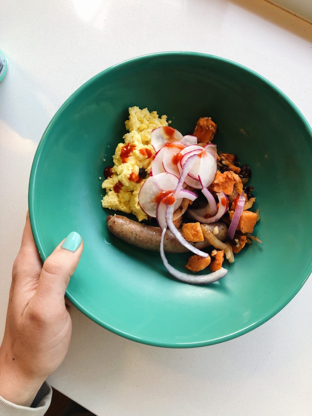 Whole foods breakfast in a green dish on a white background. In the bowl - scrambled eggs, sausage, sweet potatoes, black beans, and red onion + hot sauce