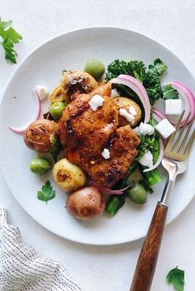 A Roasted paprika spiced chicken thigh over roasted baby potatoes and broccolini with green olives, pickled red onions, and dairy free feta cheese