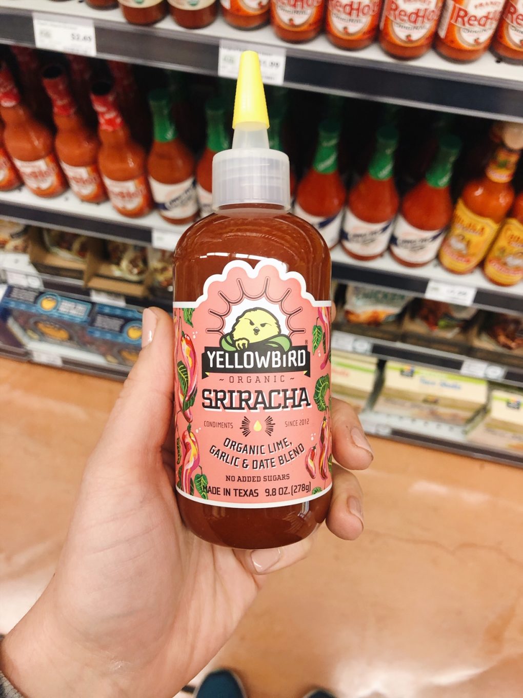Holding a bottle of yellow bird brand sriracha hot sauce in a grocery store aisle