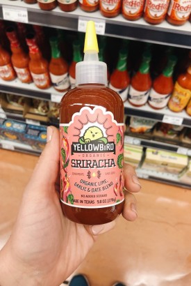 Holding a bottle of yellow bird brand sriracha hot sauce in a grocery store aisle
