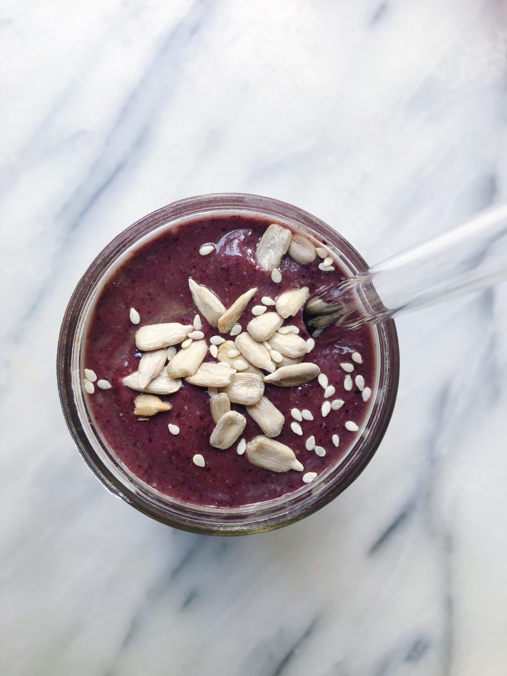  a purple berry avocado smoothie on a marble background topped with sunflower seeds and sesame seeds with a glass straw