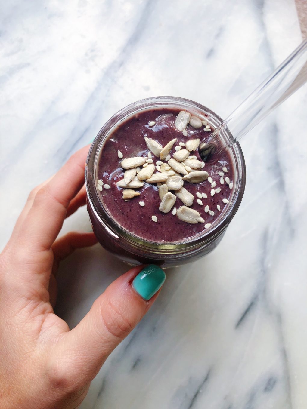 left hand holding a purple berry avocado smoothie on a marble background topped with sunflower seeds and sesame seeds with a glass straw