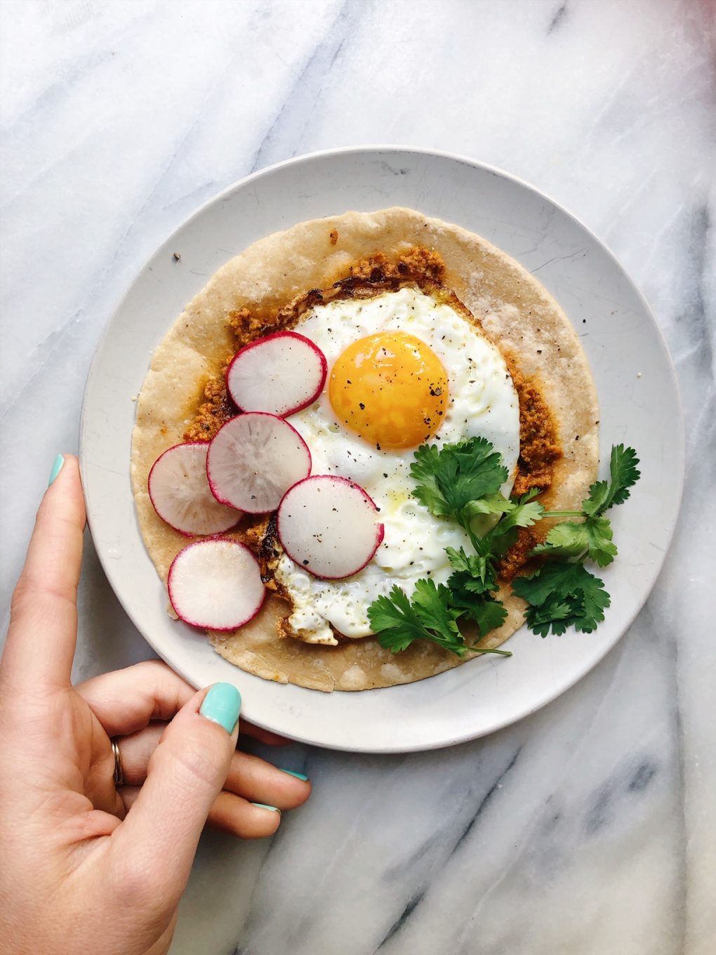 Open faced breakfast taco on a marble board and a white plate with a sunny side up egg, sliced radish, and fresh cilantro