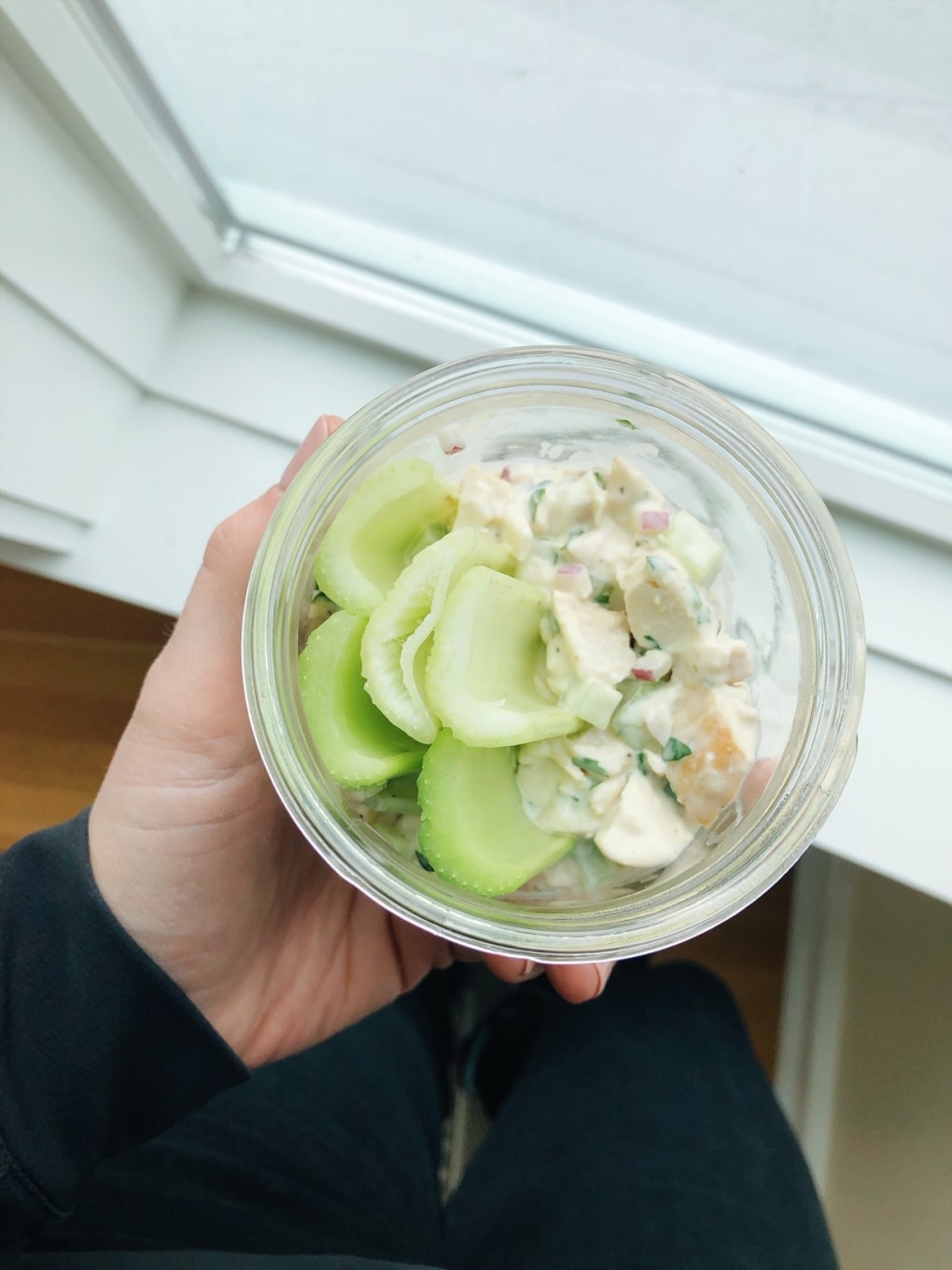 Chicken salad in a glass jar with celery sticks being held next to a window