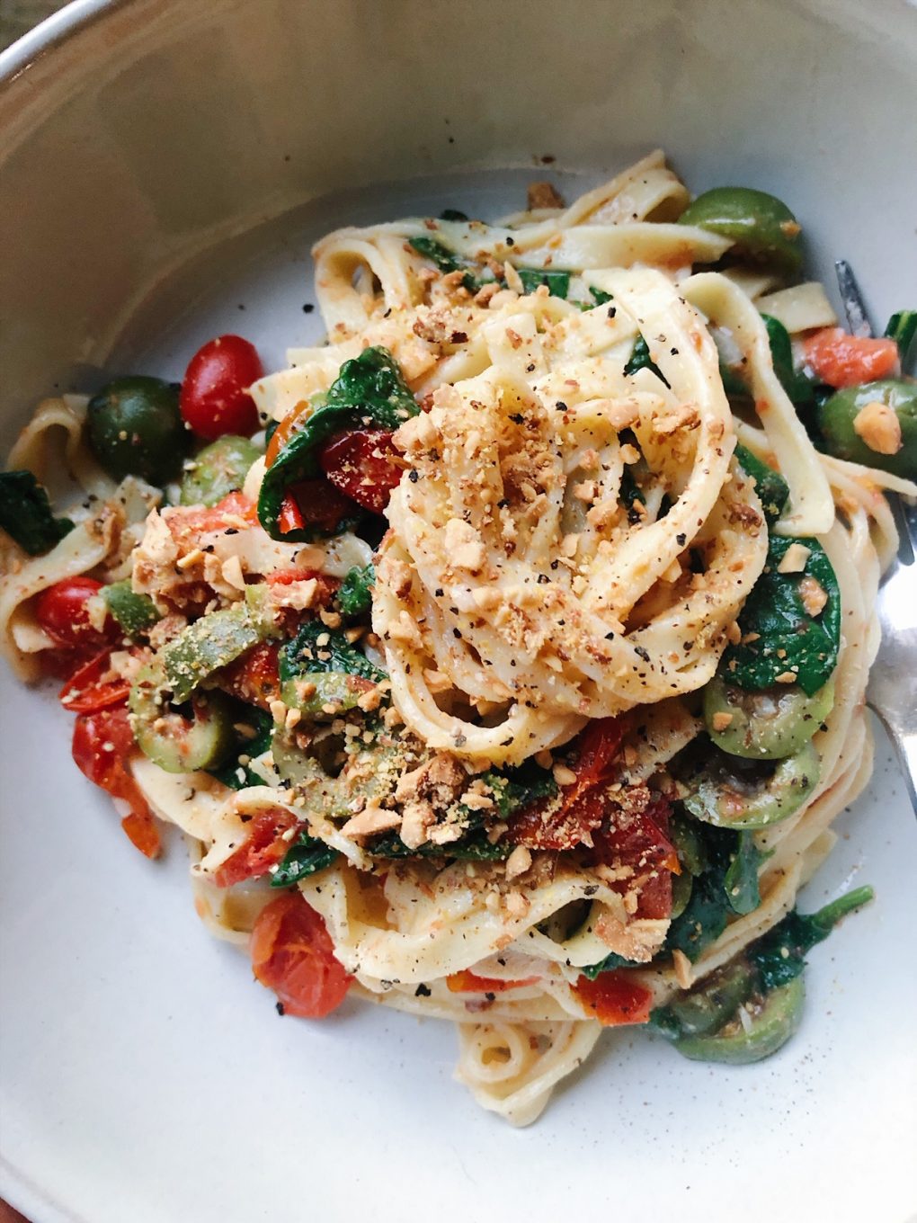 A bowl of almond flour fettuccine pasta with cherry tomatoes, spinach, and olives. Topped with toasted almond "parmesan" cheese in a big white bowl