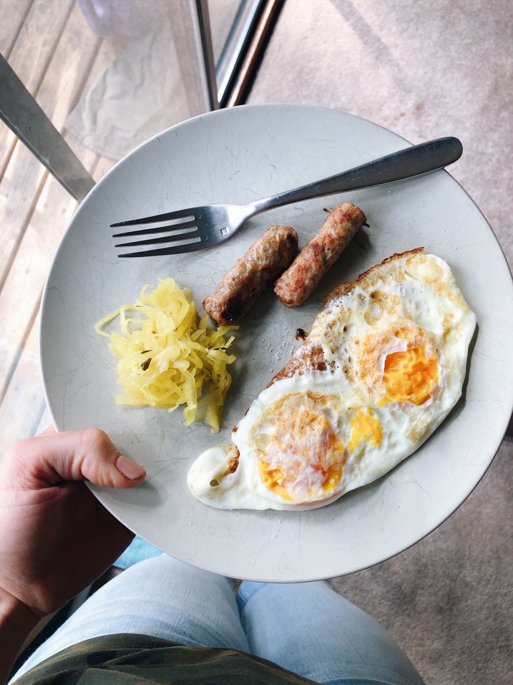 Two fried eggs with sausage and sauerkraut on a plate being held next to a window