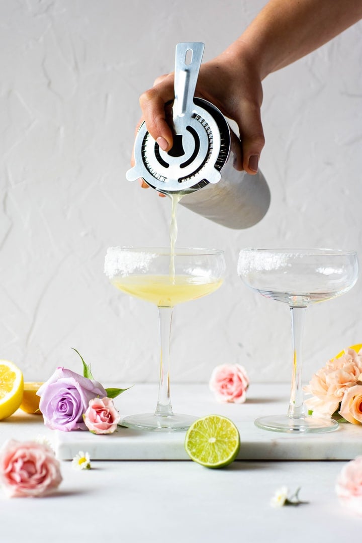 Pouring margarita into a coupe cocktail glass with a cocktail mixer surrounded by citrus and flowers