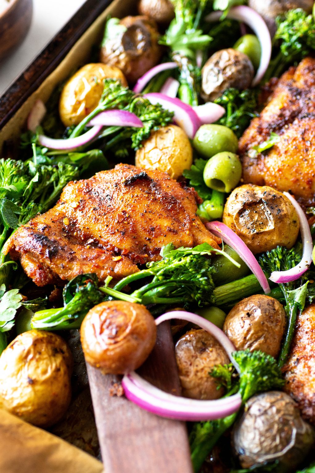 A 45 degree view of a roasting tray with spiced chicken thighs, potatos, broccolini, olives, and pickled red onions