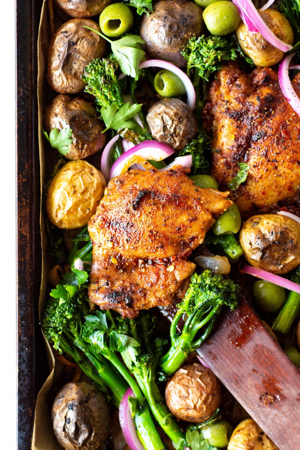 Overhead view of of a roasting tray with spiced chicken thighs, potatos, broccolini, olives, and pickled red onions