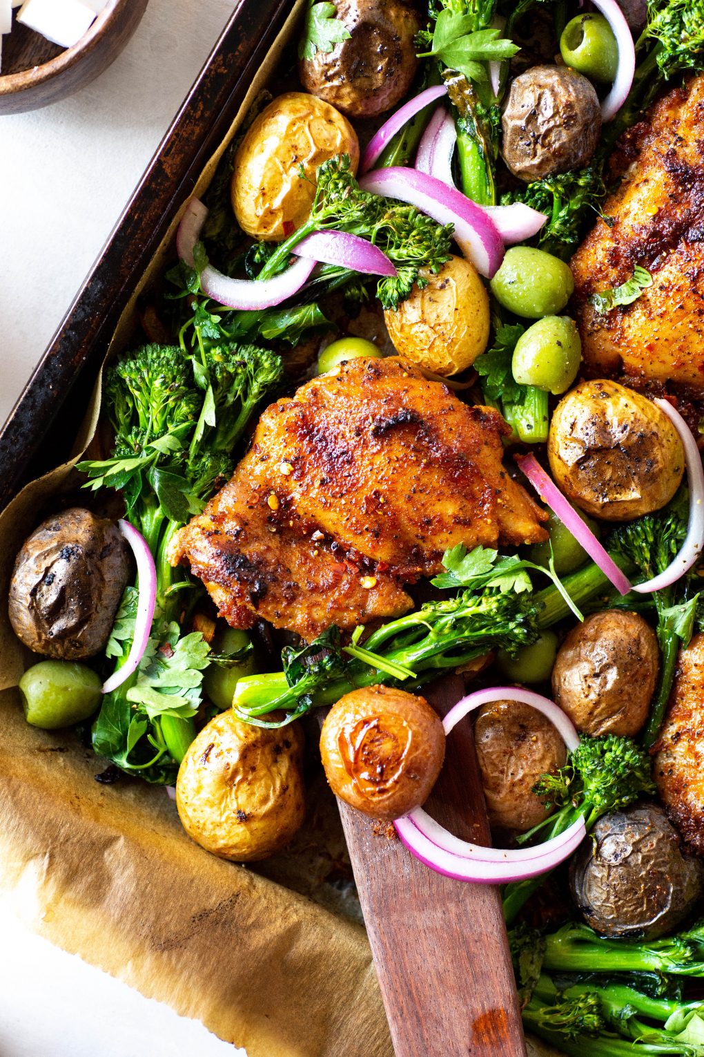 Overhead view of of an angled roasting tray with spiced chicken thighs, potatos, broccolini, olives, and pickled red onions. On a white background next to a small wooden bowl of feta cheese.