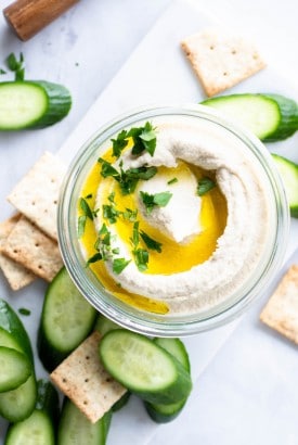 Small glass jar with swirled cauliflower hummus. Topped with olive oil and parsley, on a white background and surrounded by crackers and cucumber slices
