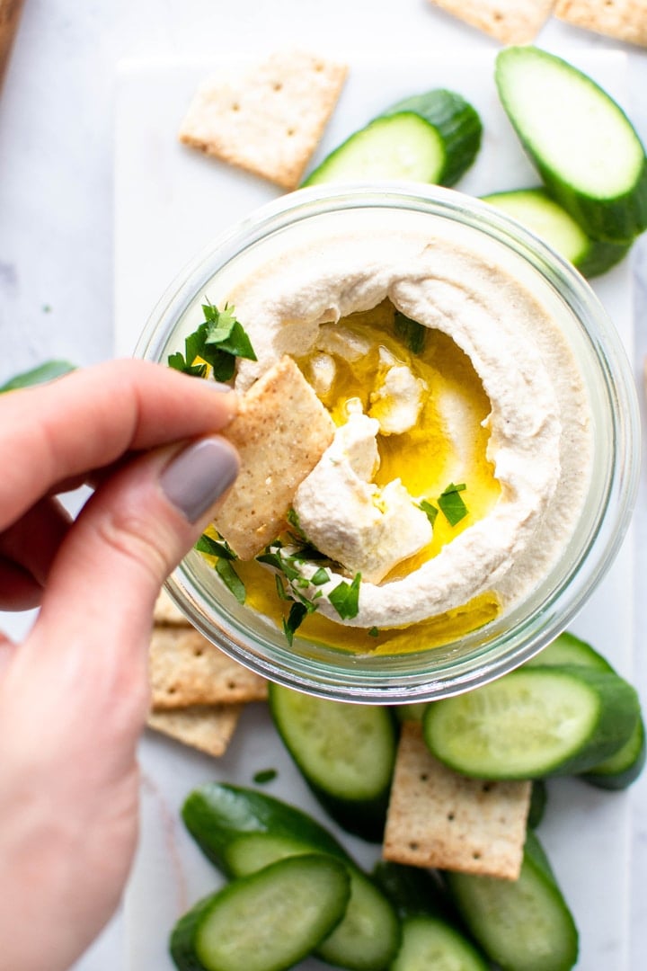 Dipping a cracker into a small glass jar with swirled cauliflower hummus. Topped with olive oil and parsley, on a white background and surrounded by crackers and cucumber slices