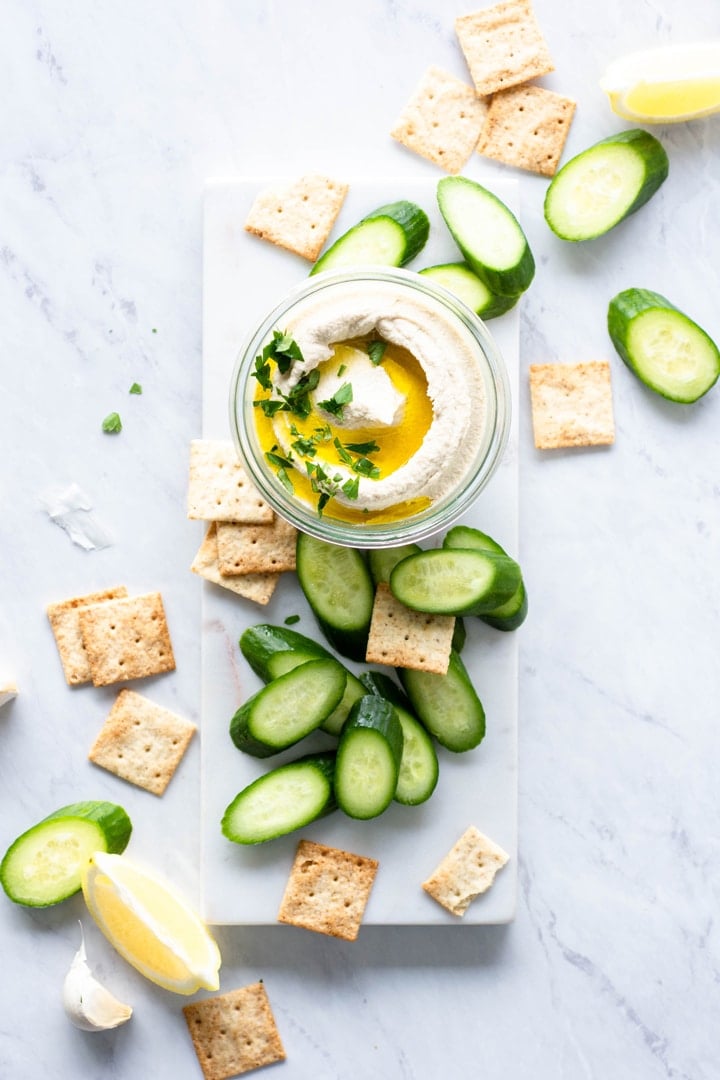 Small glass jar with swirled cauliflower hummus. Topped with olive oil and parsley, on a white background and surrounded by crackers and cucumber slices