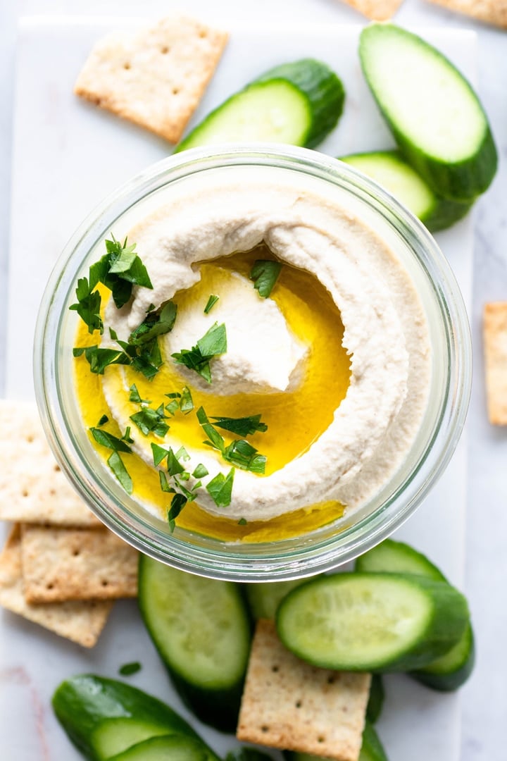 Close up view of a small glass jar with swirled cauliflower hummus. Topped with olive oil and parsley, on a white background and surrounded by crackers and cucumber slices