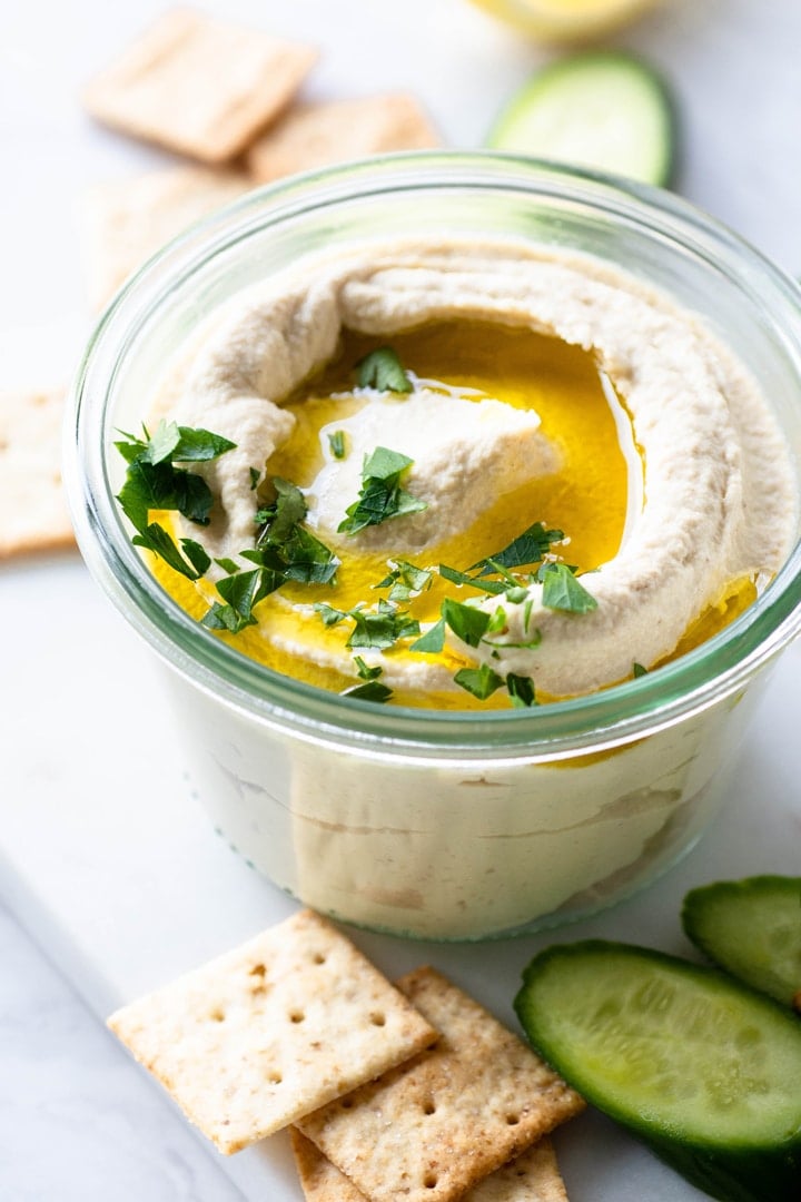 Side view of small glass jar with swirled cauliflower hummus. Topped with olive oil and parsley, on a white background and surrounded by crackers and cucumber slices