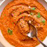 A wide, ceramic bowl of bright orange romesco sauce, garnished with chopped almonds and fresh parsley. A spoon is dipping into the center of the bowl to show the smooth yet rustic texture.