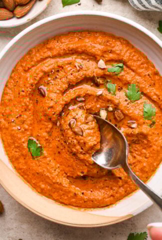 A wide, ceramic bowl of bright orange romesco sauce, garnished with chopped almonds and fresh parsley. A spoon is dipping into the center of the bowl to show the smooth yet rustic texture.