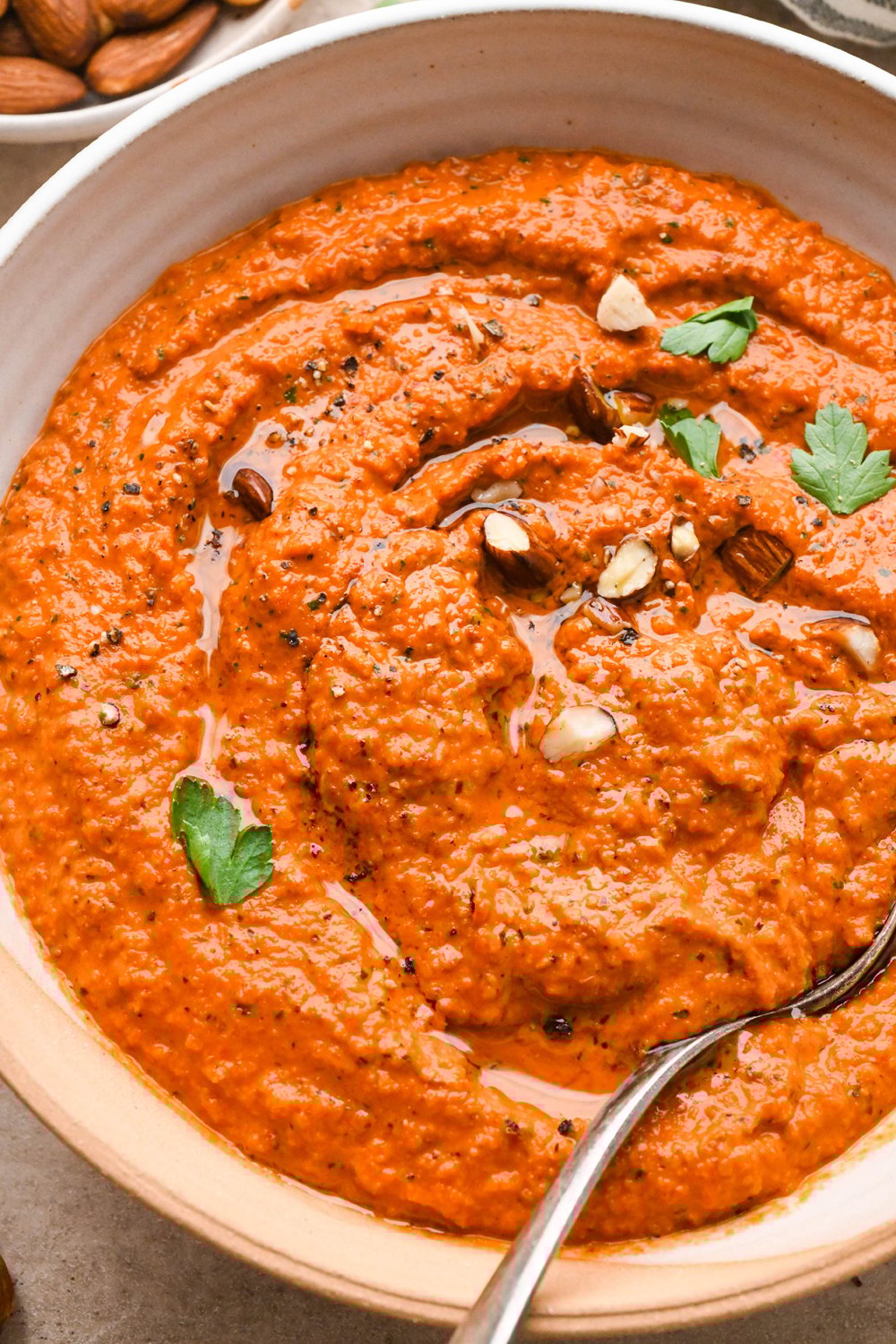 A wide, ceramic bowl of bright orange romesco sauce, garnished with chopped almonds and fresh parsley. A spoon is angled into the sauce on the side of the bowl.