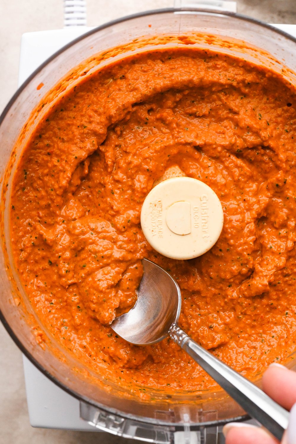 Dairy free romesco sauce in the container of a food processor, a spoon is angled into the condiment to show the rustic yet creamy texture.