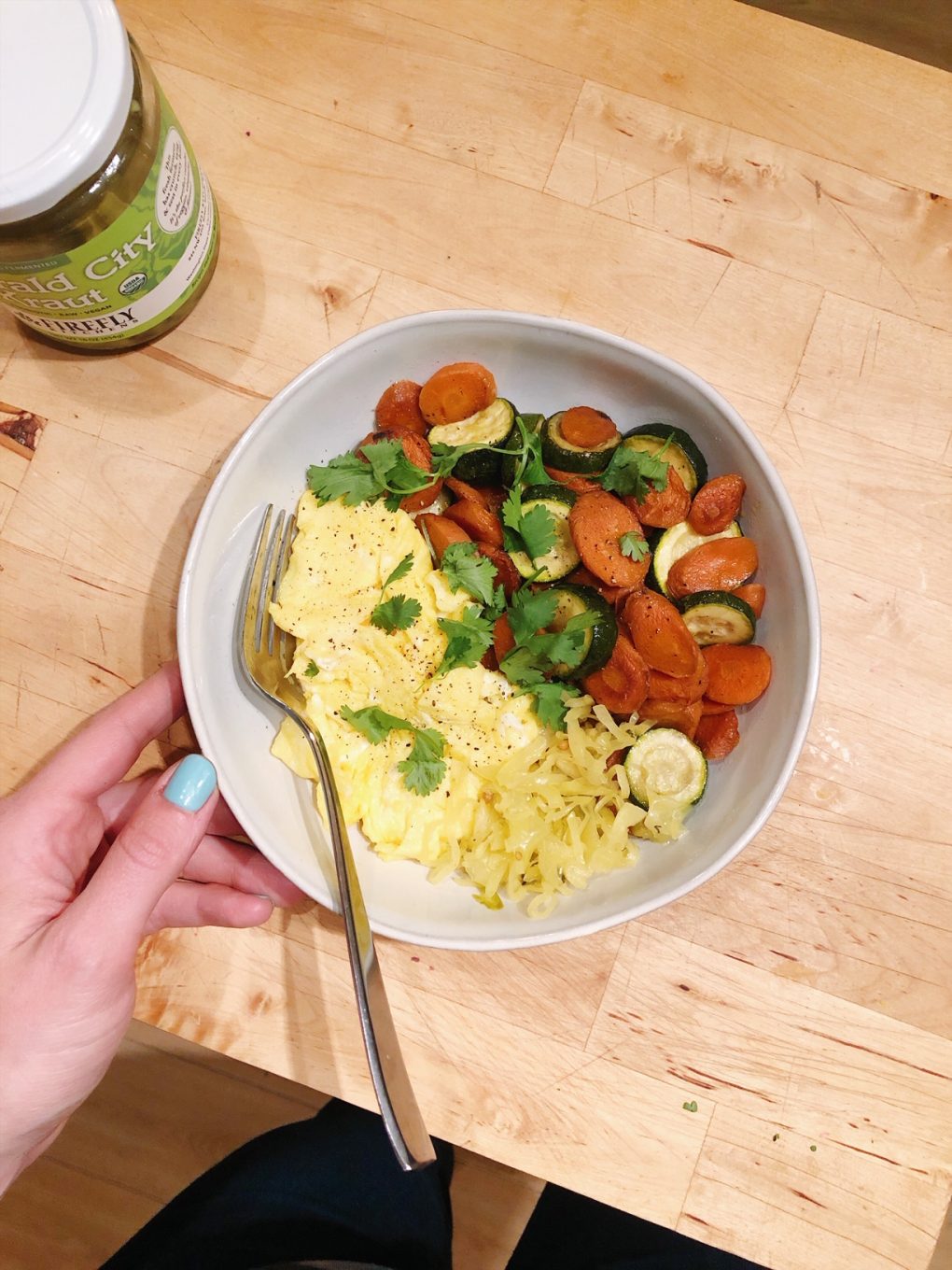 Soft scrambled eggs in a large white bowl alongside some pan roasted carrots and zuchinni with cilantro sprinkled on top and a scoop of saurkraut, over wooden kitchen counter