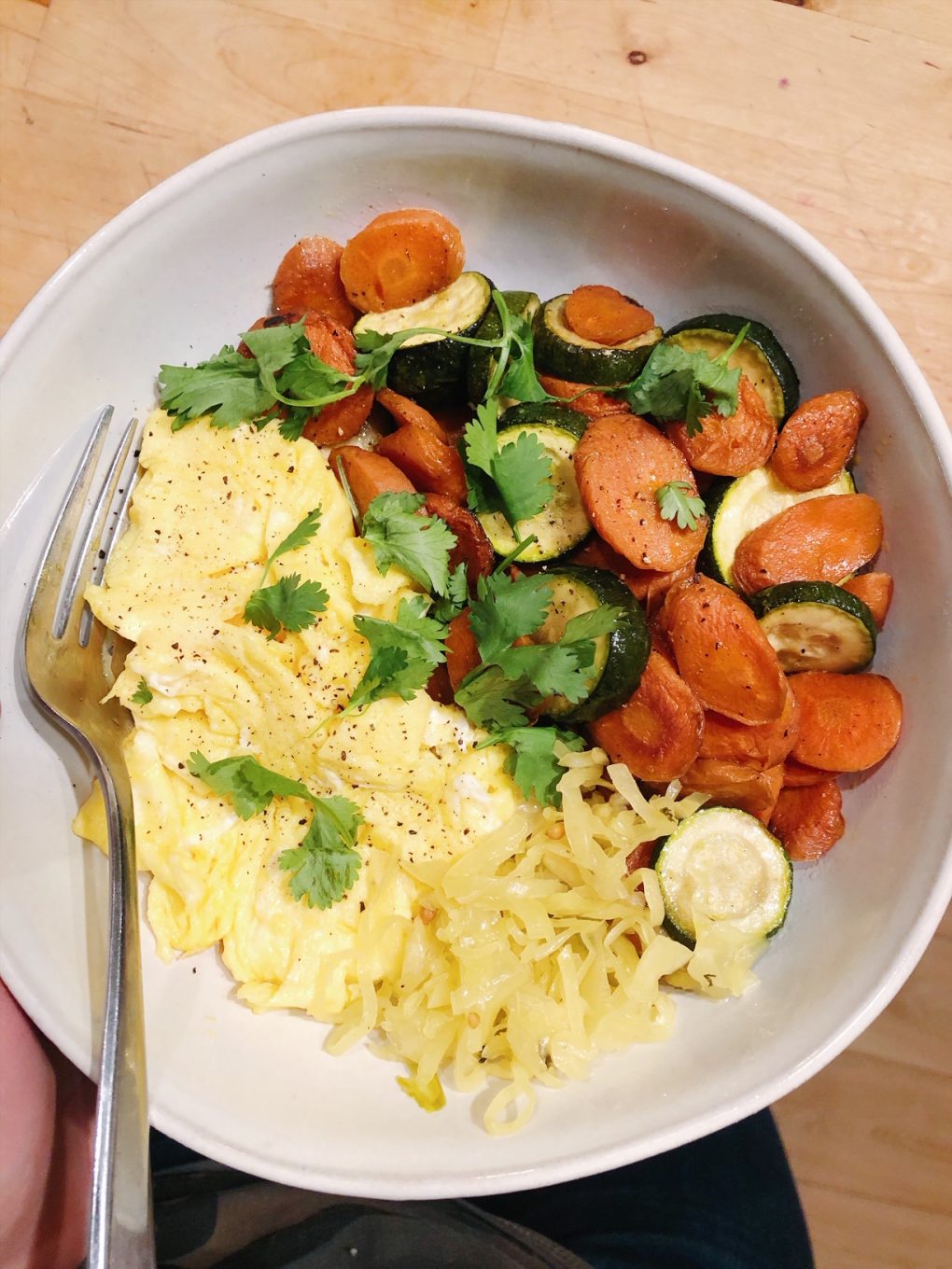Close up of soft scrambled eggs in a large white bowl alongside some pan roasted carrots and zuchinni with cilantro sprinkled on top and a scoop of saurkraut, over wooden kitchen counter
