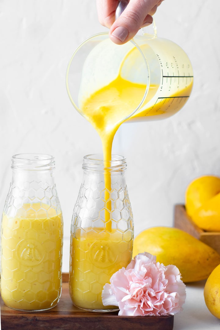 Pouring golden mango lassi into one of two side by side smoothie jars next to a pile of honey mangoes and pink carnations