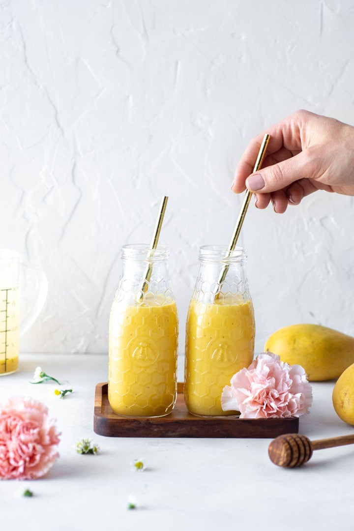 Two jars of golden hued mango lassi, side by side on a small rectangular wooden tray with large pink carnations in the frame. Hand placing a straw in one of the drinks from the right hand side of the image.
