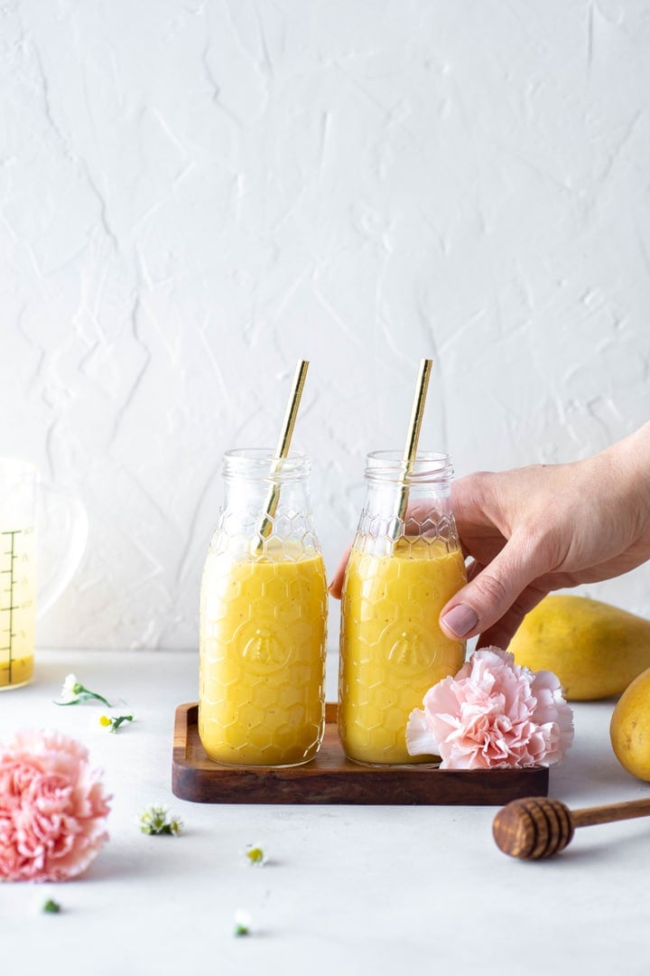 Hand in the right frame of the picture grabbing one of two side by side golden mango lassi's in smoothie jars against a white back drop