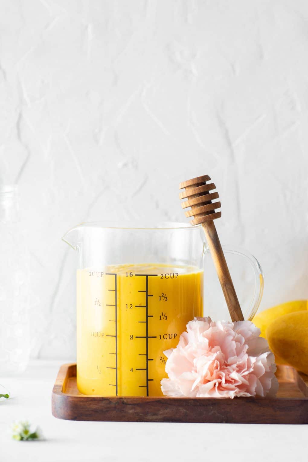 Measuring cup filled with golden mango lassi with a honey dipper leaning up against the side of the glass against a white back drop next to a pink carnation.