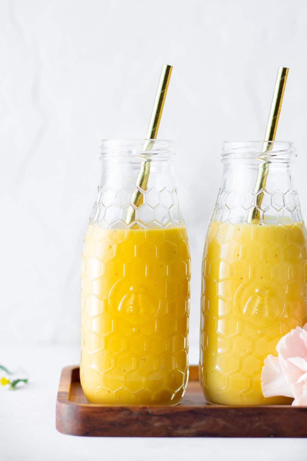Close up side view of two bottles of a golden vegan mango lassi side by side on a small wooden platter against a white background