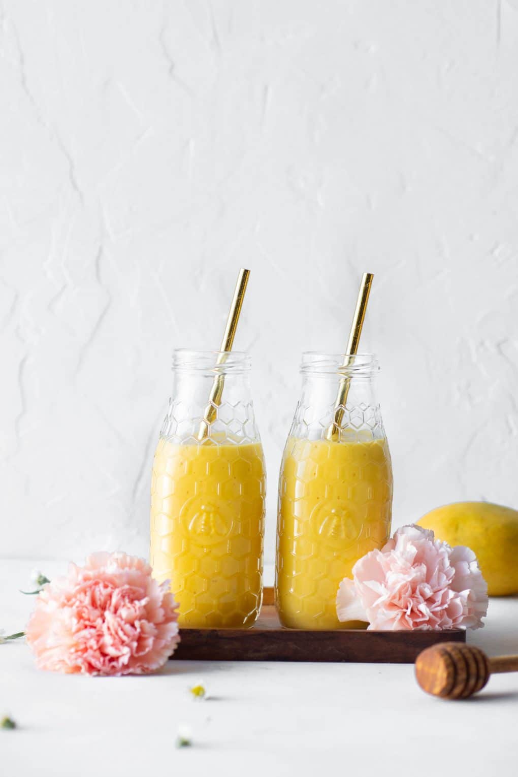 Far away side view of two bottles of a golden vegan mango lassi side by side on a small wooden platter surrounded by pink flowers against a white background
