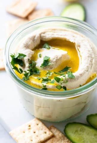 Side view of small glass jar with swirled cauliflower hummus. Topped with olive oil and parsley, on a white background and surrounded by crackers and cucumber slices