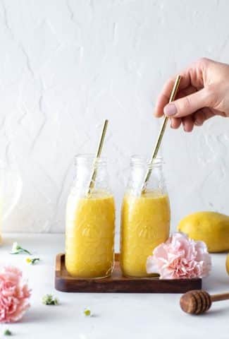 Two jars of golden hued mango lassi, side by side on a small rectangular wooden tray with large pink carnations in the frame. Hand placing a straw in one of the drinks from the right hand side of the image.