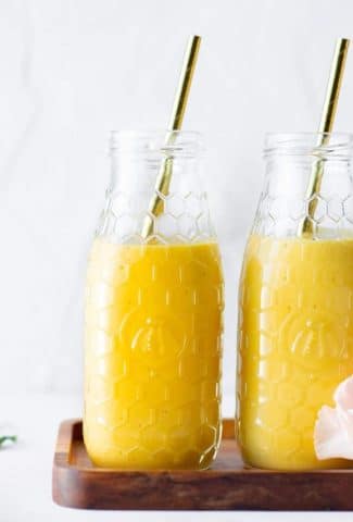 Close up side view of two bottles of a golden vegan mango lassi side by side on a small wooden platter against a white background